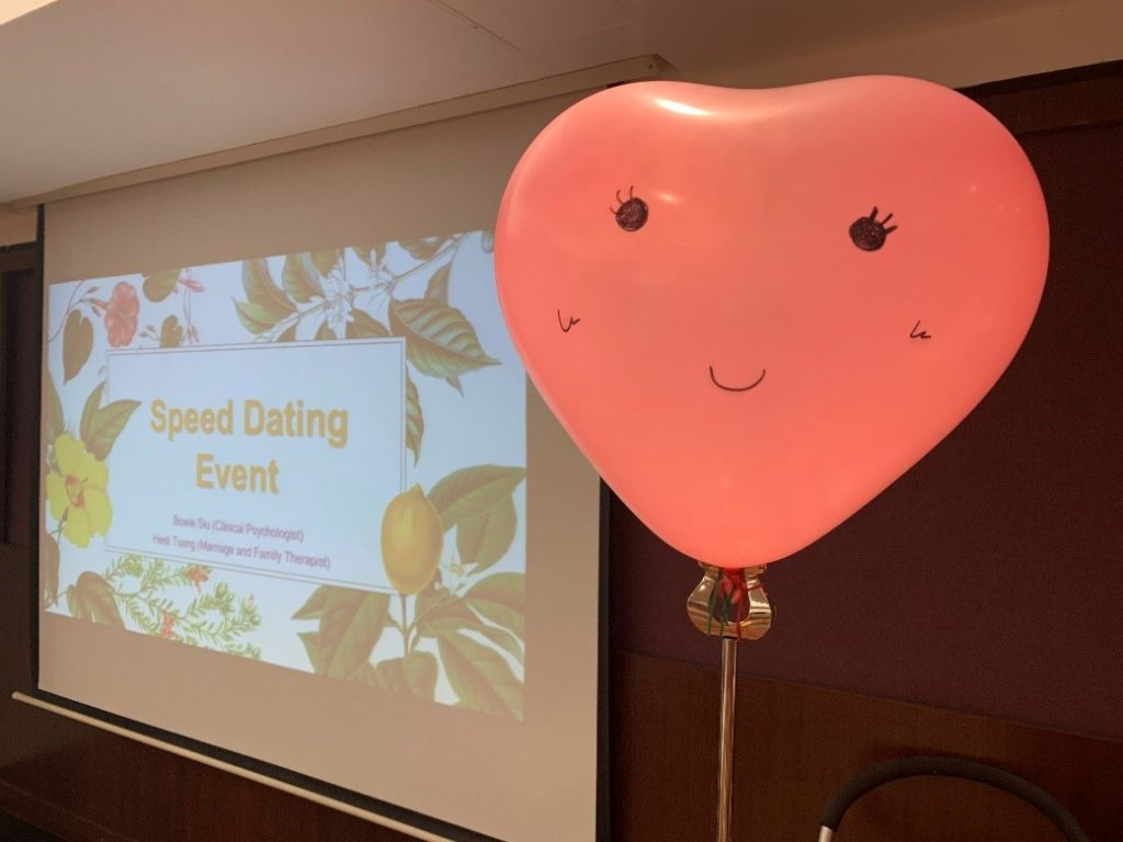 A heart-shaped balloon with a smiley face presented in front of the “Speed Dating Event” projector screen